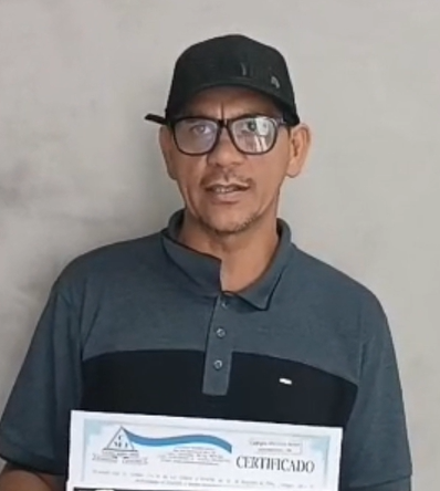 Aluno do Supletivo Sertão em Serra Talhada segurando seu certificado de conclusão do ensino médio. O aluno esta de bonê preto, camisa cinza até a parte do peito, e abaixo preto. Também esta usando óculos de vista. Aos fundo, uma parede cinza.